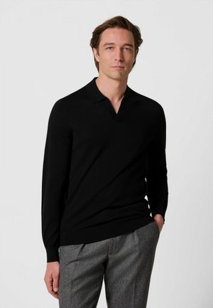 Homme aux cheveux bruns portant une chemise noire à manches longues avec col et un pantalon gris, debout avec une main dans la poche sur un fond uni.