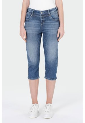 Femme portant un pantalon capri en denim bleu moyen et des baskets blanches, debout, les mains relâchées le long du corps devant un fond uni.