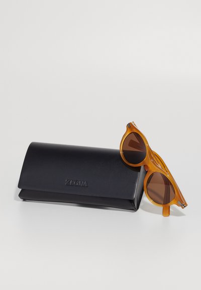 Ochelari de soare oranj cu o ramă rotunjită și lentile maro, alături de un toc din piele neagră gravat cu "ZEGNA."