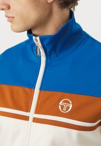 Chaqueta de pista azul y naranja con rayas blancas, cuello alto y cierre de cremallera. Presenta un logotipo circular en el pecho. Textura de tela suave.