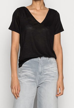 Kobieta ubrana w czarny krótki top z dekoltem w serek, włożony do wysokich, szerokich spodni w jasnoszarym kolorze, stojąca na jednolitym tle.