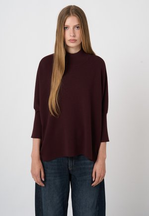 Jonge vrouw met lang, recht blond haar, gekleed in een losse donkere bordeauxrode trui en blauwe jeans, staand tegen een effen lichte achtergrond.