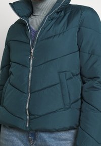 Turkis puffjakke med chevron-vattering, stående krave og sølvlynlås. Har en frontlomme og en tekstureret strikket rullekrave under.