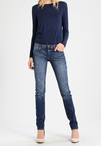 Top azul marino de manga larga con lunares blancos, combinado con vaqueros ajustados azules. Modelo con tacones rosa claro. Conjunto informal de diseño contemporáneo.