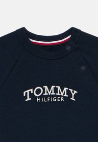 Navy modra felpa s okroglim izrezom, ki vsebuje izvezeno logotip "TOMMY HILFIGER". Spodnji izrez ima črtast akcent v rdeči, beli in modri barvi.