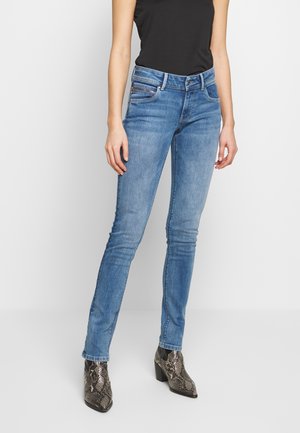Vrouw draagt aansluitende blauwe jeans, zwart mouwloos topje en bruine enkellaarsjes met slangenhuidpatroon, staand tegen een effen witte achtergrond.