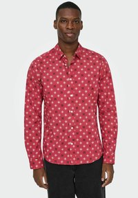 Man in een rood overhemd met witte en zwarte sterrenpatronen, gecombineerd met zwarte broek, staande tegen een neutrale achtergrond.