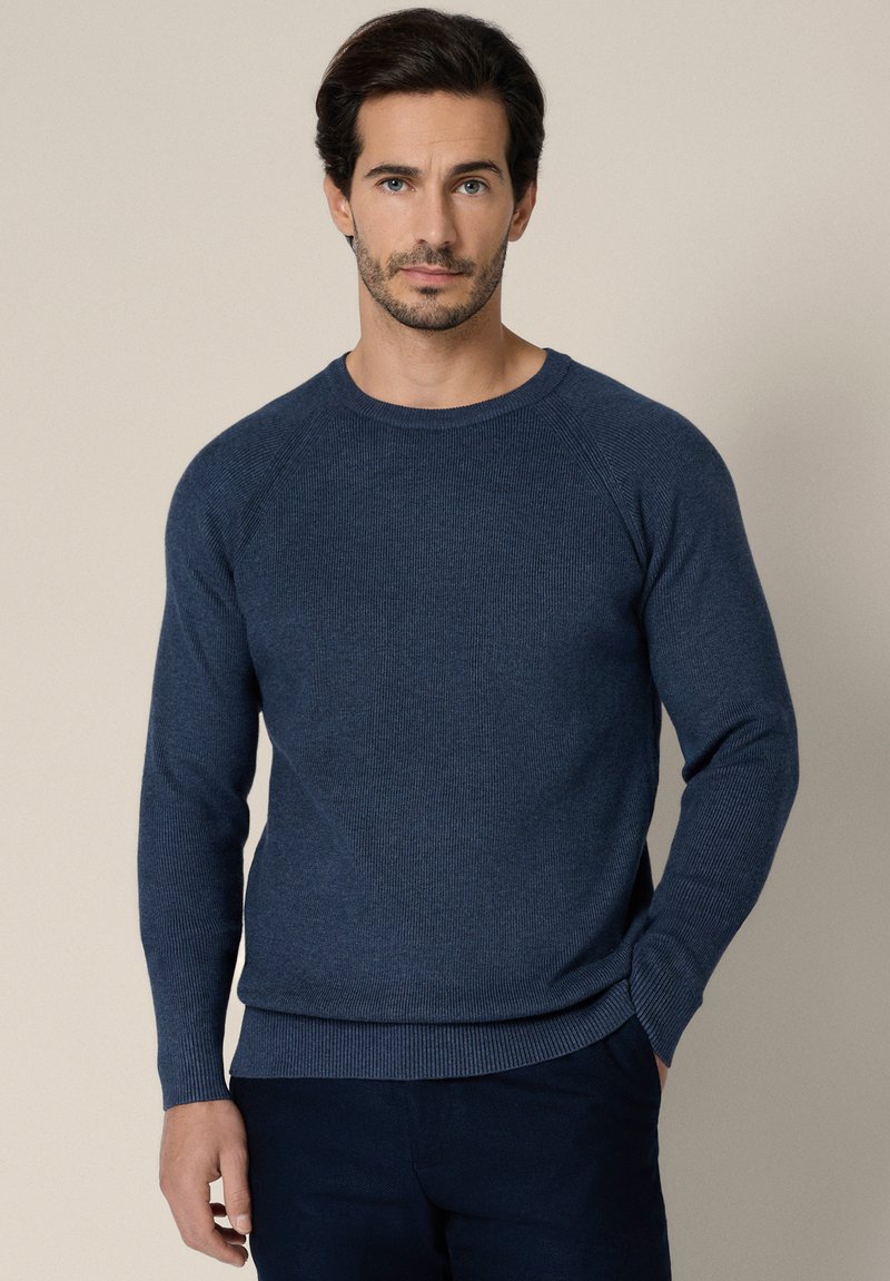 Mężczyzna o ciemnych włosach, ubrany w granatowy sweter z długim rękawem i ciemne spodnie, stojący na tle jednolitego beżowego tła.