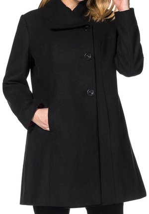 Femme portant un manteau noir boutonné à col montant, main gauche dans la poche et main droite près du visage, cheveux blonds visibles.