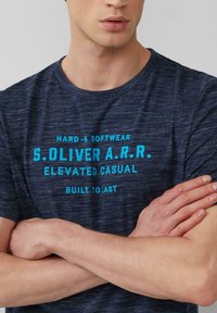 Donkerblauwe T-shirt met een gemêleerde textuur, voorzien van turquoise tekst: "HARD & SOFTWEAR S.Oliver A.R.R. ELEVATED CASUAL BUILT TO LAST."