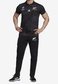 Svart pikétröja med ormbunke-mönster och vita logotyper, i kombination med svarta byxor och svarta sneakers. Materialet verkar lätt och texturerat.