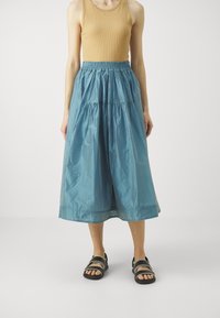 Gonna blu chiaro lungo il ginocchio in un tessuto liscio e leggero con vita elastica e dettagli arricciati. Indossata con un top beige e sandali.