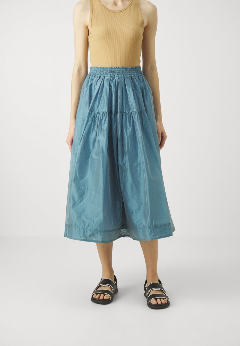 Gonna blu chiaro lungo il ginocchio in un tessuto liscio e leggero con vita elastica e dettagli arricciati. Indossata con un top beige e sandali.