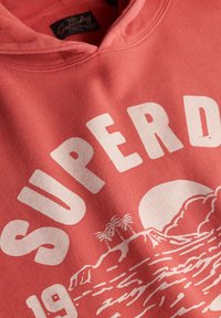 Sudadera color coral con texto blanco "SUPER" y gráfico de playa. Cuenta con capucha con cordón y textura suave. Tonos terrosos con motivos isleños.