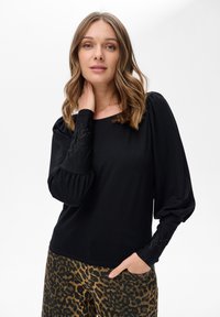 Kobieta z falującymi, jasnobrązowymi włosami, ubrana w czarny top z długim rękawem z haftowanymi mankietami oraz spodnie w panterkę, pozująca z ręką na szyi.