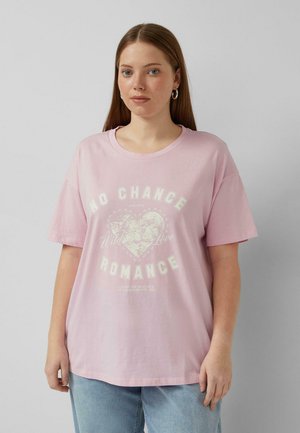 Frau mit glatten braunen Haaren, die Creolen und ein lockeres pinkfarbenes T-Shirt mit der Aufschrift "No Chance Romance" und einem Herzgrafik trägt.