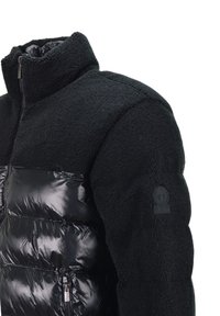 Giacca in pile nera e giacca a sacco lucida con colletto alto e chiusura con zip. Presenta un tessuto testurizzato e un patch con logo sulla manica.