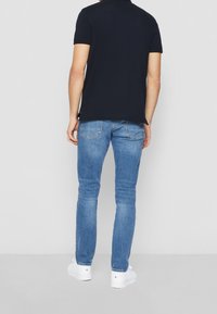 Man som står med ryggen mot kameran iförd en svart kortärmad skjorta, ljusblå jeans och vita sneakers på en neutral bakgrund.