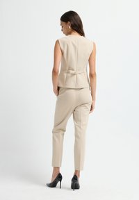Blazer sans manches beige au design ajusté, avec un détail au dos et un fermoir doré. Associé à un pantalon slim beige et des talons noirs.