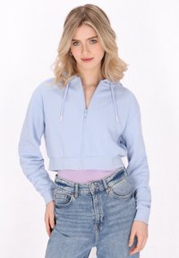 Felpa azzurra corta con zip, cappuccio, polsini a costine e una texture liscia, abbinata a jeans in denim a vita alta.