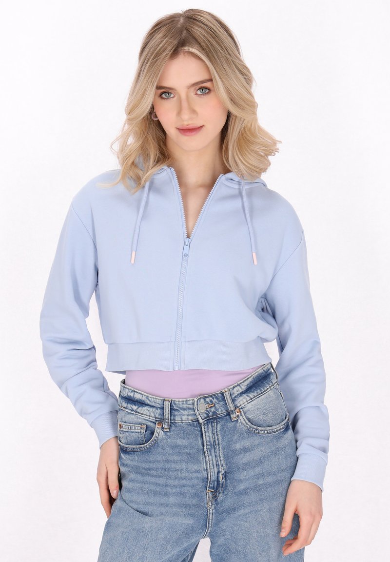 Felpa azzurra corta con zip, cappuccio, polsini a costine e una texture liscia, abbinata a jeans in denim a vita alta.
