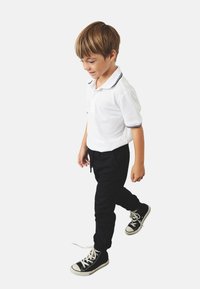 Polo bianco a maniche corte con rifiniture nere, abbinato a pantaloni neri aderenti e sneakers alte nere. Bambino che cammina, sorridendo leggermente.