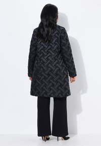 Svart quiltad kappa med ett texturerat vävt mönster, långa ärmar och knä-längd design, kombinerad med svarta vida byxor och klackskor.