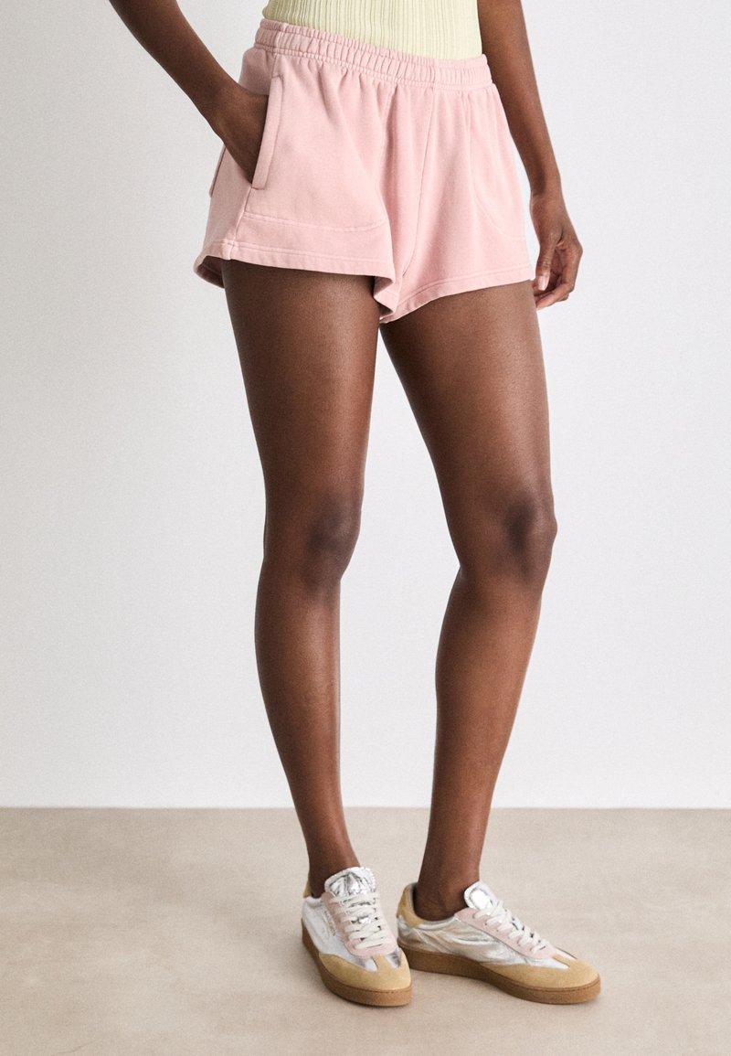 Shorts en coton rose clair avec une taille élastique, deux poches latérales et une coupe décontractée, associés à des baskets argentées et beige.