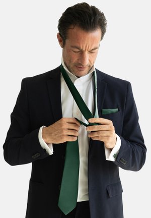 Corbata verde con un acabado texturizado, llevada con una camisa blanca y un traje oscuro. Un pañuelo de bolsillo en un tono verde a juego es visible.