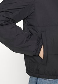 Veste noire avec poignets élastiques et une main insérée dans la poche latérale, portée sur un jean en denim bleu.