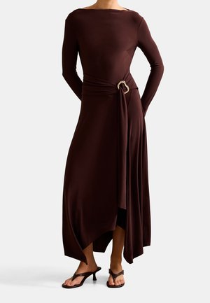 Frau trägt ein langärmeliges, dunkelbraunes asymmetrisches Kleid mit einer dekorativen Gürtelschnalle und schwarzen Sandalen mit Absatz.
