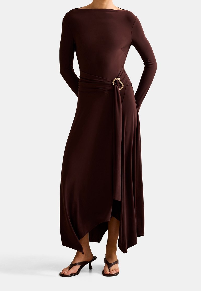 Femme portant une robe asymétrique marron foncé à manches longues avec une boucle de ceinture décorative et des sandales à talons noires.
