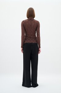 Maglia lunga marrone a maniche lunghe con collo alto, abbinata a pantaloni neri a gamba larga. La maglia ha un design a coste, leggermente trasparente.