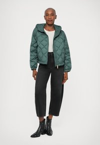 Kvinne står iført en turkise quilted jakke, hvit skjorte, svarte cropped bredbukser og svarte ankelstøvler med metallhull.