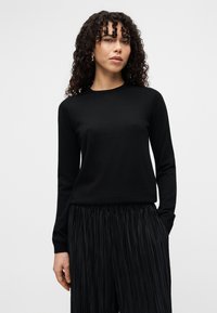 Μαύρο πουλόβερ με μακριά μανίκια, κατασκευασμένο από απαλό υλικό με στρογγυλή λαιμόκοψη, σε συνδυασμό με μαύρο πλισέ παντελόνι, δημιουργώντας μια ομαλή αντίθεση υφής.