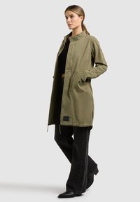 Žena stojaca v dlhej olivovo zelenej bunde, čiernom krátkom topu, čiernych širokých nohaviciach a čiernych špicatých čižmách na jednoduchom sivom pozadí.