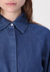 Camisa de ganga com colarinho pontudo, fecho de botão e mangas raglan. O tecido é de um azul médio com um acabamento texturizado.