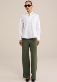 Camicia bianca con bottoni e maniche lunghe, abbinata a pantaloni verdi oliva larghi e occhiali da sole neri a forma quadrata. Scarpe nere piatte.