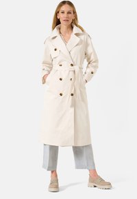 Trench beige court avec de gros boutons ronds, taille ceinturée, grand col et poches latérales ; associé à un pantalon court gris clair.