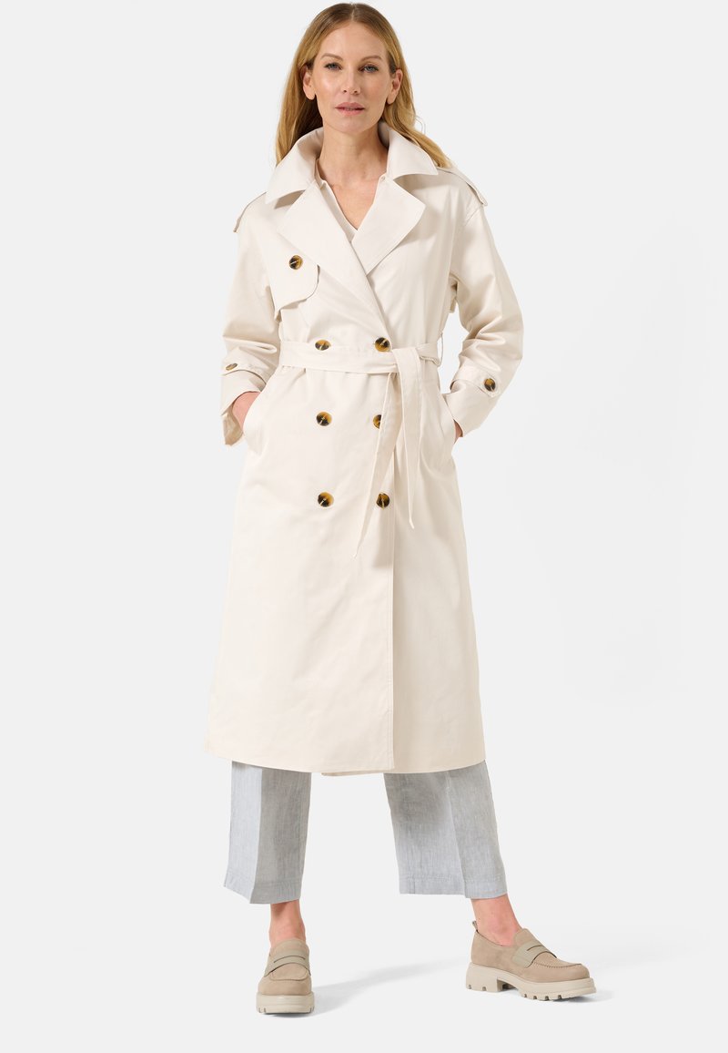 Trench beige court avec de gros boutons ronds, taille ceinturée, grand col et poches latérales ; associé à un pantalon court gris clair.