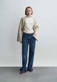 Μπεζ μάλλινη ζακέτα πάνω από ένα ανοιχτόχρωμο τοπ, συνδυασμένη με μπλε τζιν παντελόνι με φαρδύ πόδι και σκουρό κόκκινα παπούτσια. Απλός σχεδιασμός, υφασμάτινη υφή και άνετη εφαρμογή.