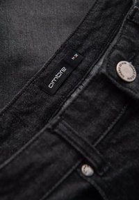 Mustat denim-farkut, joissa on sileä rakenne, varustettuna hopeisella napilla ja "ombre"-tunnisteella. Sisältää ompeluyksityiskohtia.