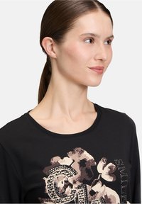 Vrouw met bruin haar dat vastgebonden is, die een zwart shirt met een beige bloemen- en studdecoratie draagt, en iets naar rechts kijkt.