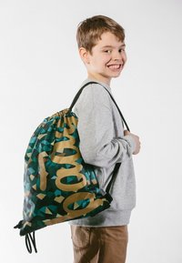 Zelený a modrý geometrický vzorovaný taška na stahovací šňůrku se zlatým nápisem, vyrobená z látky, s černými popruhy a staženým vrchním uzávěrem.