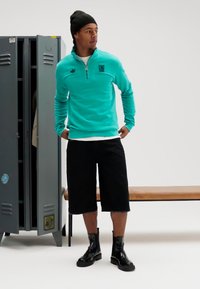 Πλεκτή μπλούζα με φερμουάρ σε τιρκουάζ χρώμα, μαύρα σορτς μέχρι το γόνατο και μαύρες γυαλιστερές μπότες. Το φόντο περιλαμβάνει μια γκρι ντουλάπα και ένα ξύλινο παγκάκι.