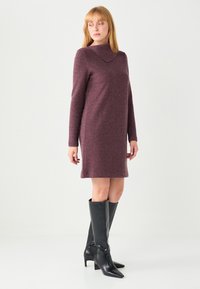Robe bordeaux à manches longues avec un col arrondi, longueur mi-cuisse, associée à des bottes en cuir noires montantes jusqu'aux genoux avec des bouts pointus.