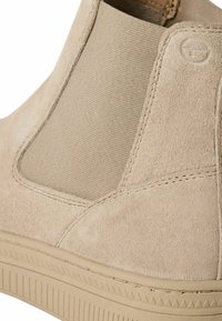 Bottines en suède beige avec panneaux latéraux élastiques et semelle en caoutchouc texturée. Comprend des coutures surpiquées et un petit logo sur le côté.
