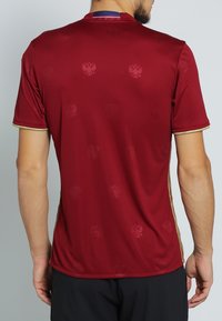 Röd idrottströja med subtila präglade mönster, gyllene detaljer vid kragen och ärmarna, samt en slank, jämn textur.