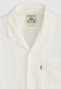 Fehér, rövid ujjú ing, finom textúrális mintával, klasszikus gallérral, gombos záródással, és egy mellzsebbel, amelyen egy piros Levi's címke található.