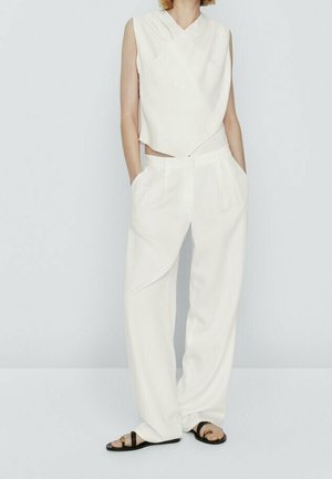 Woman wearing sleeveless white draped top and matching wide-leg white trousers with black flat sandals, hands in pockets, standing against a plain background.